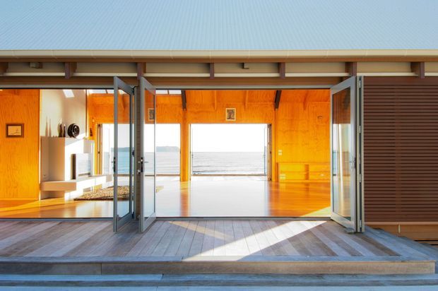 Bright contemporary coastal-facing interior opening onto a wide timber deck, with warm orange timber wall panelling, polished timber flooring, large bifold glazed doors, and direct views toward the sea. The open-plan room appears airy and strongly connected to the waterfront exterior.