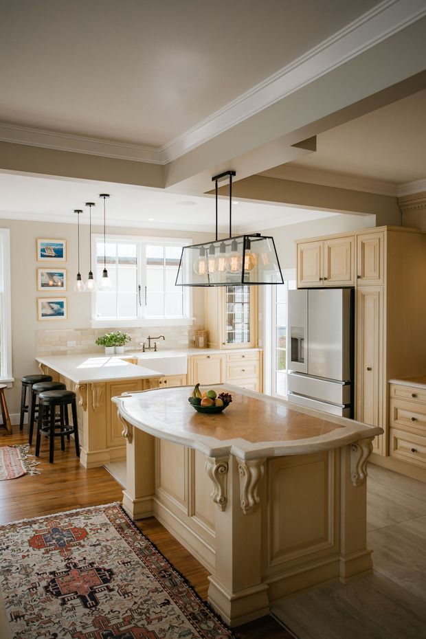 Bright, traditional-style kitchen with extensive cream-painted cabinetry, a large ornate central island with a light stone benchtop, stainless-steel refrigerator, white-framed windows, pendant lighting, timber flooring, and a patterned area rug. Decorative cornices, classical trim, and carved island brackets add a refined character.