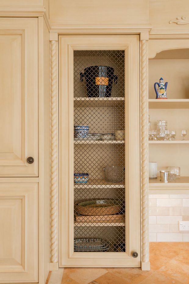 Close-up of a traditional built-in kitchen storage cabinet with cream-painted paneled cabinetry, twisted rope-style trim, and a glazed wire-mesh door. Shelves display crockery and glassware, with open display shelving and a tiled backsplash visible to the right. The floor appears to be warm terracotta-toned tile.