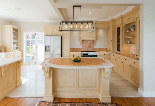 Bright, spacious traditional kitchen with ornate cream-painted cabinetry, a large decorative island with carved corbels, paneled rangehood surround, open shelving, stone benchtops, and a brick feature backsplash around the stove. A large black metal lantern pendant hangs above the island, with French doors bringing in natural light. Timber flooring transitions to pale kitchen tile, and a patterned rug adds colour in the foreground.