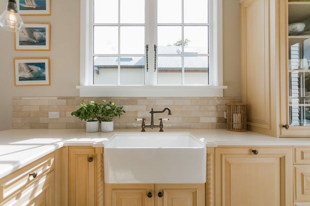 Bright, traditional-style kitchen focused on a deep white farmhouse apron-front sink beneath a multi-pane window. Warm honey-toned timber cabinetry, a beige rectangular tiled splashback, dark metal bridge-style tapware, open shelving, and framed artwork create a refined, welcoming fit-out.