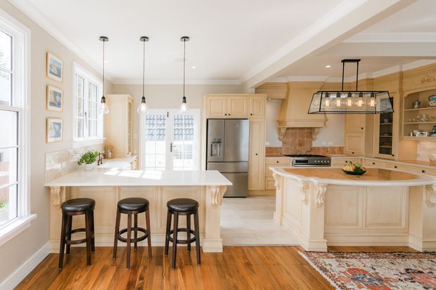 Bright, spacious traditional kitchen with cream painted cabinetry, ornate island detailing, white stone benchtops, stainless-steel appliances, pendant lighting, and warm timber flooring. The room includes a large central island with three stools, a second sculpted island or peninsula, extensive glazing, and decorative architectural trim.