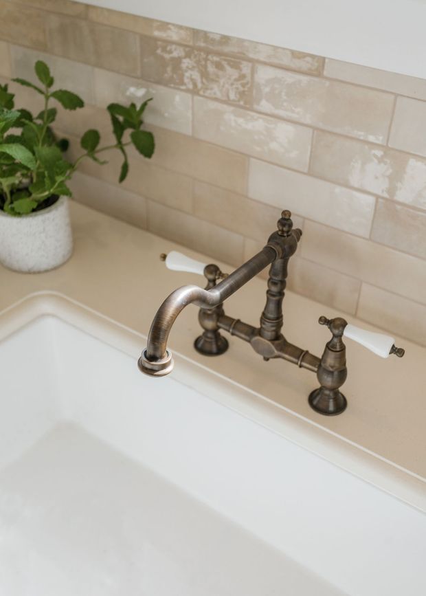 Close-up of a freestanding white bathtub with a vintage-style dark bronze bridge faucet and white ceramic lever handles. The tub is set against a glossy cream-beige rectangular tiled wall, with a small potted plant on the ledge.