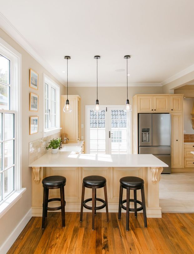 Bright, traditional-style kitchen featuring a large cream-painted island with a white benchtop, decorative corbels, and seating for three on dark wooden stools. The room has warm timber flooring, cream cabinetry, stainless-steel appliances, pendant lighting, framed artwork, and abundant natural light from multiple windows and glazed French doors.