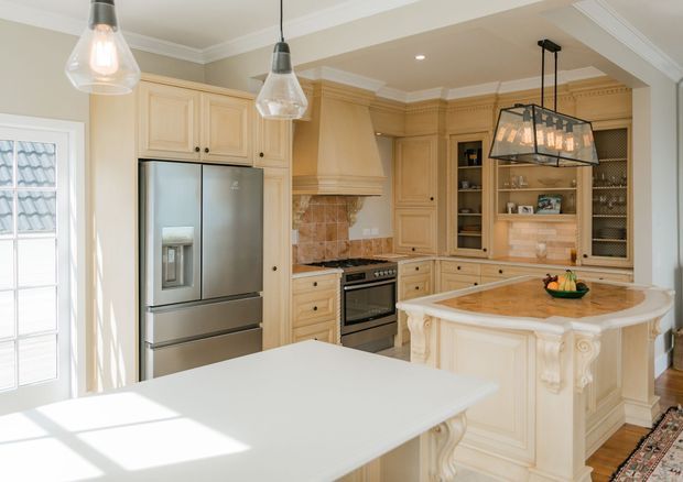 Bright, traditionally styled kitchen with cream painted cabinetry, detailed crown moulding, carved island corbels, stainless-steel refrigerator and range, stone backsplash, timber flooring, and pendant lighting. A large central island and foreground work surface dominate the room.