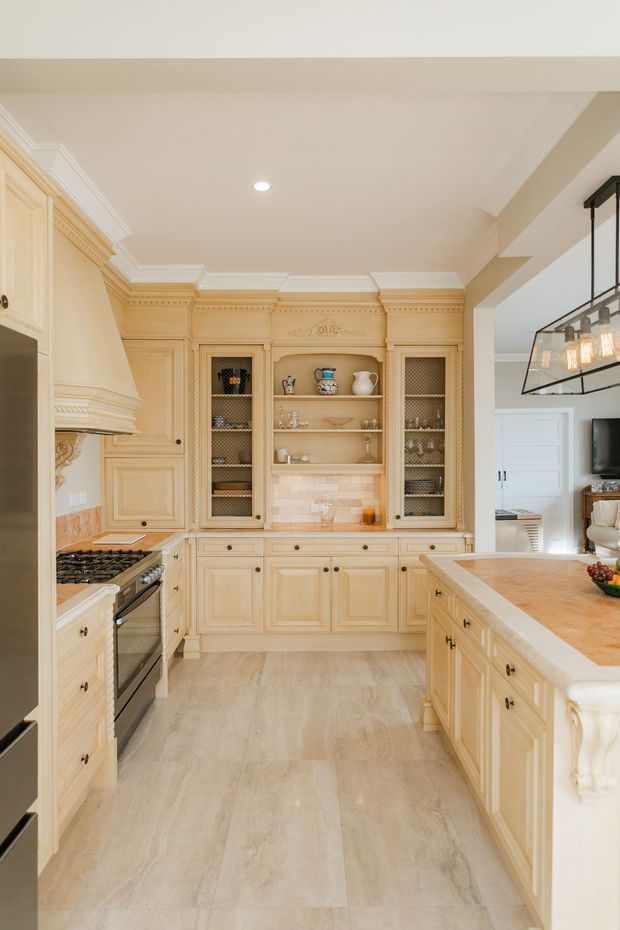 A warm, traditional kitchen featuring extensive cream-painted raised-panel cabinetry, ornate crown moulding and decorative cornice details, glass-front display cabinets, a tiled backsplash, a large central island, and a stainless-steel range. Light stone-look flooring and beige walls create a refined, cohesive palette. An adjacent living area is visible beyond the kitchen.