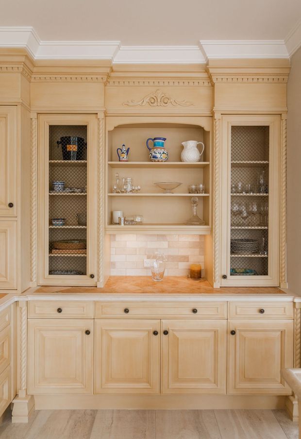 A detailed traditional built-in display and storage cabinet, likely within a kitchen or dining area. The pale timber cabinetry features raised-panel doors, decorative mouldings, carved detailing, antique-style dark knobs, glass-fronted side cupboards with diamond-pattern mesh, and open central shelving displaying ceramics and glassware. A warm beige subway-tile backsplash and timber benchtop sit above pale stone or timber-look flooring.