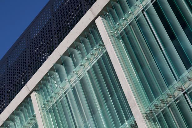 Angled close-up of a modern commercial building facade, featuring tall turquoise-tinted vertical glass fins, pale concrete structural frames, and a dark perforated metal screen along the upper section against a clear blue sky.