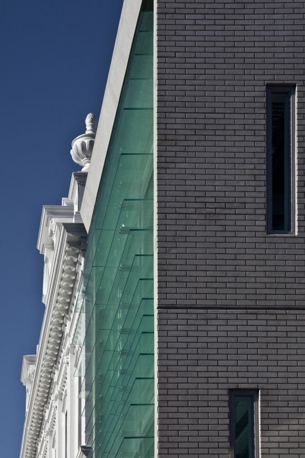 Close-up exterior architectural photograph showing a contemporary dark brick facade with narrow vertical windows beside a large green-tinted glazed wall. A highly ornate white historic facade with classical cornices and decorative detailing is visible immediately adjacent, creating a strong contrast between heritage and modern construction. Clear blue sky forms the background.