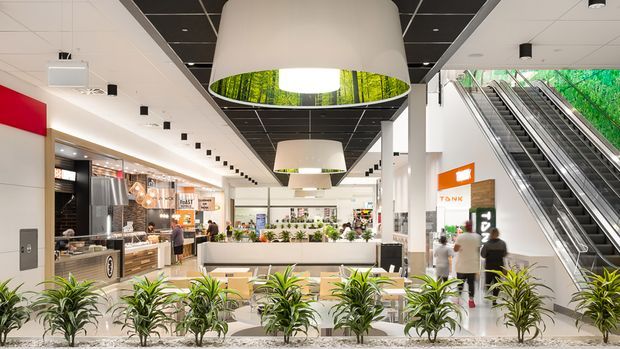 Bright contemporary commercial interior resembling a shopping-centre or retail dining concourse. The open atrium contains multiple food and retail tenancies, a central planter-lined seating area, escalators, extensive white finishes, black ceiling panels, and large drum pendant lights with illuminated forest imagery. Green planting forms a prominent foreground divider.