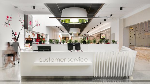 Bright contemporary commercial customer service reception area with a large white curved-front desk, integrated illuminated signage, computer workstations, and a sculptural vertical-slat counter extension. The space features polished light flooring, white walls, a black acoustic ceiling grid with oversized pendant lights, recessed lighting, indoor plants, and retail or service areas extending behind the desk.
