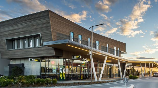 Exterior view of a contemporary multi-storey commercial building with a dark horizontal timber-clad upper level, extensive ground-floor glazing, projecting metal awnings, and distinctive angled white support columns. Retail tenancies occupy the lower level, with landscaped frontage and a covered pedestrian edge. The image is photographed at sunset under a partly cloudy sky.