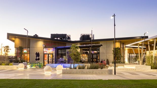 Twilight exterior view of a contemporary low-rise commercial building or retail precinct. The facade combines warm wood-toned cladding, pale rendered or panelled walls, extensive glazing, illuminated shopfronts, and a projecting covered entry or terrace. A broad paved forecourt includes integrated planters, sculptural spherical lights, seating, young trees, and landscaped lawn in the foreground.