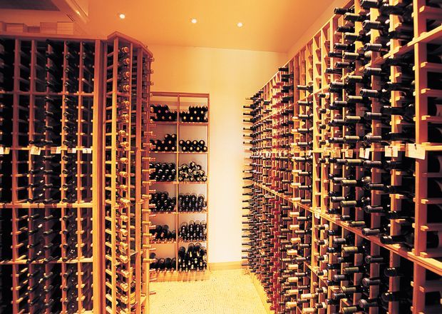 A dedicated wine storage room lined with extensive floor-to-ceiling timber wine racks on both sides and shelving at the rear. Hundreds of bottles are stored horizontally in an aisle layout, with warm recessed ceiling lighting and a pale tiled floor.