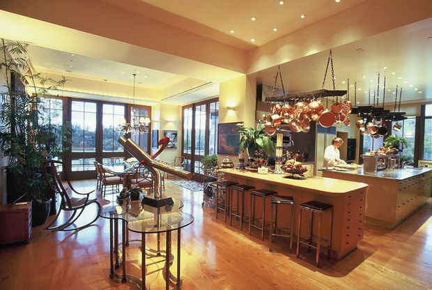 A warm, spacious open-plan interior combining a kitchen and living or dining area. The kitchen features a large island with seating, extensive timber cabinetry, stainless-steel appliances, and a prominent hanging rack displaying copper cookware. The adjacent lounge area has timber flooring, dark-framed windows, a glass coffee table, rocking chairs, plants, and abundant natural light. Recessed ceiling lighting and pendant fixtures add a welcoming atmosphere.