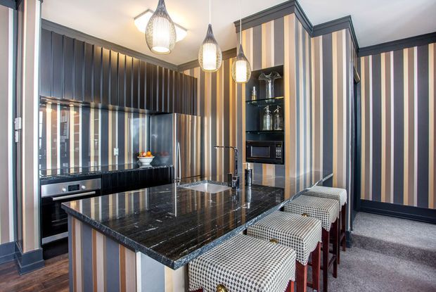 A striking, compact kitchen with dramatic vertically striped wall finishes and cabinetry in dark charcoal, taupe, cream, and muted blue-grey tones. The room features a large polished dark stone island with integrated sink, patterned upholstered bar stools, black built-in cabinetry, glass display shelving, and three sculptural pendant lights. The overall design is bold, highly coordinated, and strongly graphic.