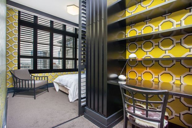 A compact bedroom and adjoining study nook divided by a dark, slatted partition. The bedroom has a low bed with white bedding, a black metal lounge chair, carpeted flooring, and large windows with dark plantation shutters. The study nook features built-in dark shelving, a desk, a chair, and bold yellow-and-white geometric hexagonal wallpaper. The space has a playful, high-contrast contemporary interior.