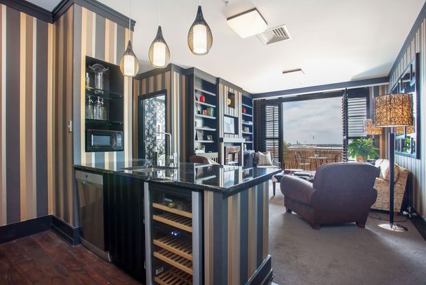 A stylish open-plan kitchen and living area with a dark island and integrated wine storage, black cabinetry, reflective benchtops, and bold vertical striped wall panelling. Pendant lights hang above the island, while a seating area with an armchair and floor lamp sits beside large louvred windows overlooking an exterior deck or balcony.