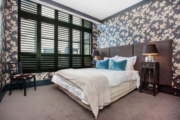 A stylish bedroom with a large upholstered bed, white bedding, turquoise accent cushions, matching bedside tables and lamps, dark floral wallpaper, black plantation shutters, and wall-to-wall carpeting. The room has a bold, refined, high-contrast decorative scheme.