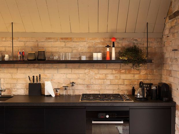 Compact industrial-style kitchen with exposed warm brick walls, dark lower cabinetry and benchtops, a gas cooktop and built-in oven, open wall shelving with glassware and accessories, and a pale timber-lined ceiling. The space has warm, subdued lighting and a strong material contrast between brick, black joinery, and timber.