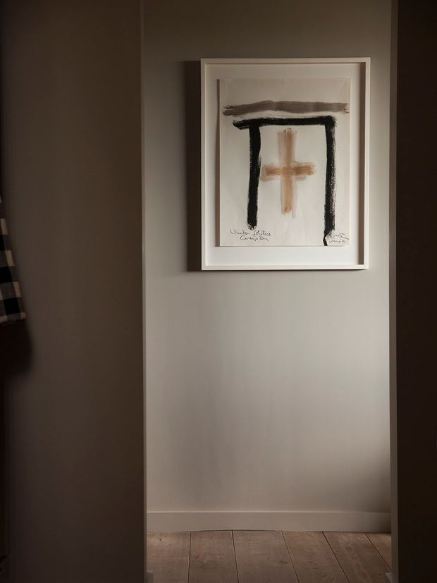 A dimly lit, narrow interior alcove or hallway with warm grey painted walls, white skirting boards, and natural timber floorboards. A single white-framed abstract artwork featuring bold black and ochre brushstrokes hangs centrally on the wall. Dark openings or adjacent spaces frame the view on both sides, creating a calm, intimate atmosphere.