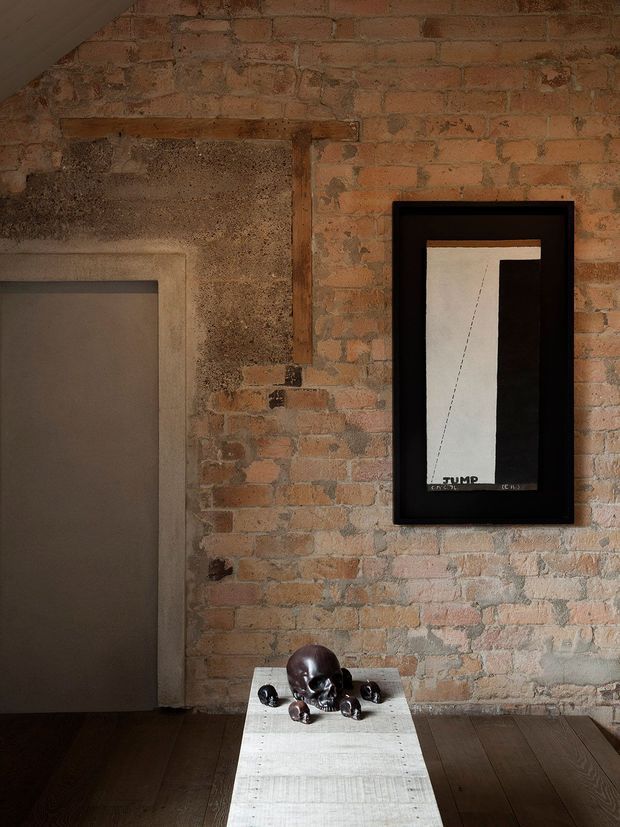 A moody, industrial-style interior detail featuring an exposed aged brick wall, a dark timber floor, and a grey recessed doorway. A framed monochrome artwork hangs on the brick wall, while a narrow white console table displays a dark sculptural object and small decorative pieces. The space has warm, subdued lighting and raw architectural character.