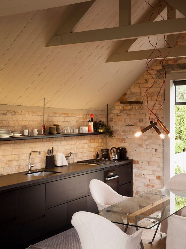 A compact loft-style kitchen and dining area beneath a steep sloping ceiling. The room features exposed pale brick walls, a white painted timber-lined ceiling with exposed beams, matte dark cabinetry, dark benchtops, open shelving, integrated appliances, and a glass dining table with curved white chairs. A sculptural hanging light with exposed red wiring adds an industrial accent, while a large window brings in natural light.