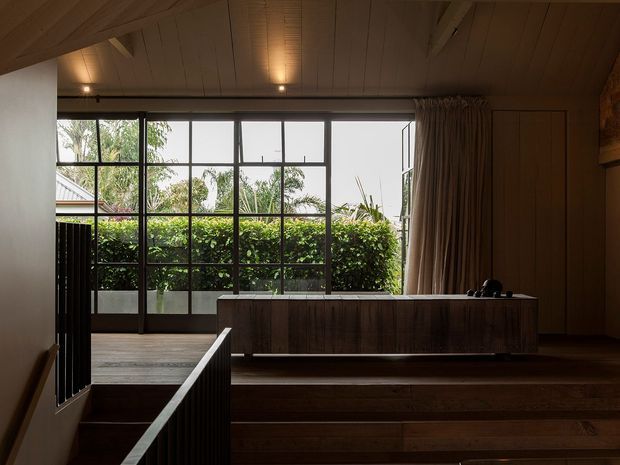 A dark, timber-lined interior landing or living area with broad stepped timber flooring, a long built-in timber cabinet, and floor-to-ceiling black-framed glazing opening toward a planted exterior. A curtain wall occupies the right side, while a stair or balustraded level change leads down in the foreground. The ceiling features warm timber boards and recessed spotlights, creating a moody, refined atmosphere.