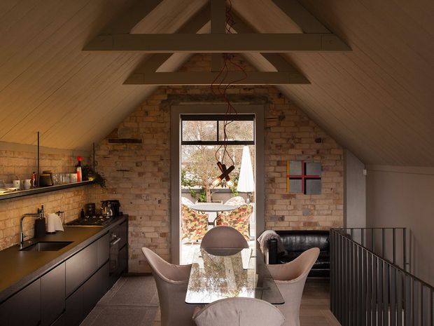 Compact attic dining and kitchenette space with a steep pitched, timber-lined ceiling and exposed dark timber trusses. A warm exposed-brick feature wall surrounds a glazed door, while dark built-in cabinetry and a black benchtop run along the left side. A glass dining table with upholstered chairs occupies the centre, and a black metal balustrade borders the stair opening on the right. The space has a warm, moody, industrial-contemporary character.