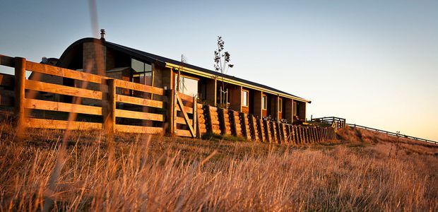 A low, elongated rural house viewed from the exterior at sunset, set among dry grasses behind a timber post-and-rail fence. The home has dark corrugated-looking pitched roof forms, warm timber cladding and exposed timber verandah elements, with a brick chimney and a long row of windows and doors opening onto a deck. The image has a warm, rustic architectural character and appears to be a single-storey dwelling.