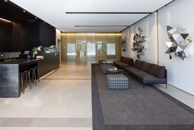 A spacious contemporary lobby or open-plan reception area with a black stone bar and stools on the left, a long dark sectional sofa and plaid ottomans on a large dark rug, polished pale tiled flooring, and sculptural geometric wall art. Full-height glazing and warm concealed lighting create a refined, high-end atmosphere.