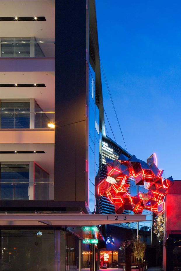 Vertical evening view of a contemporary multi-storey commercial building and pedestrian frontage. The facade combines dark metal cladding, extensive glazing, balconies with glass balustrades, and strong linear lighting. A large, vivid red geometric illuminated art installation projects over the entrance canopy, creating a dramatic urban streetscape.