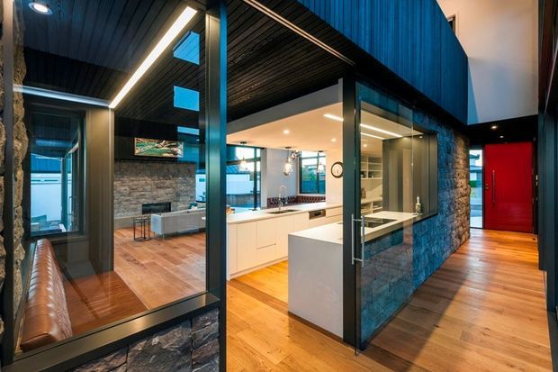 Interior view of a contemporary open-plan kitchen and living area. The space features a white kitchen with a central island, extensive glazing, warm timber flooring, dark timber-lined ceilings, black-framed glass partitions and doors, and a prominent rough stone-clad wall. A vivid red entry door provides a strong accent. The image also shows a reflected living area with a fireplace and seating.