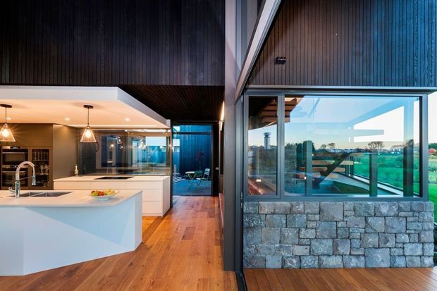 A contemporary open-plan kitchen viewed from an adjoining interior space, featuring a large white angular island, white fixed cabinetry, pendant lighting, timber flooring, dark vertical timber wall and ceiling lining, and extensive glazing. A stone-clad lower wall and landscaped surroundings are visible through the glazing.