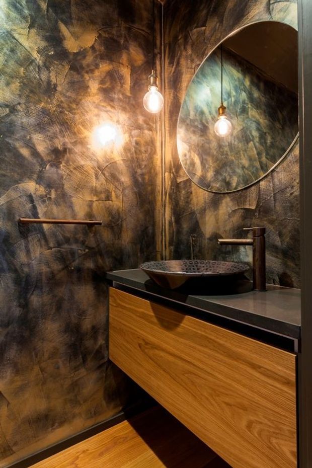 A compact, dramatically styled bathroom with a floating timber vanity, dark countertop, vessel basin, bronze-toned wall-mounted tapware, large round mirror, and exposed pendant bulbs. The walls have a richly mottled metallic bronze and charcoal textured finish, creating a moody, luxurious atmosphere.