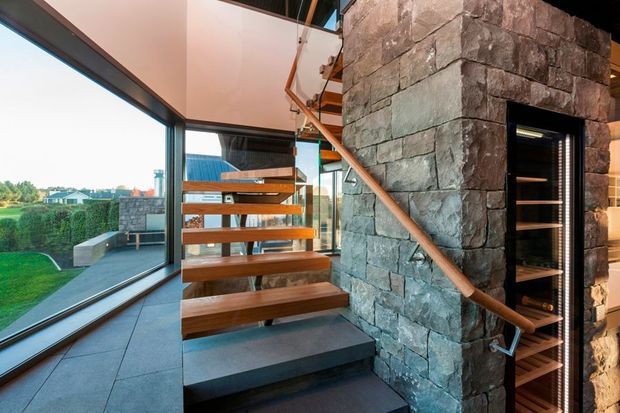 Interior view of a contemporary stairway beside a substantial rough-stone feature wall. The staircase has open-riser timber treads with a slim timber handrail and dark grey structural elements, while full-height glazing brings in views of the landscaped exterior. A dark-framed built-in shelving or storage niche is integrated into the stone wall.