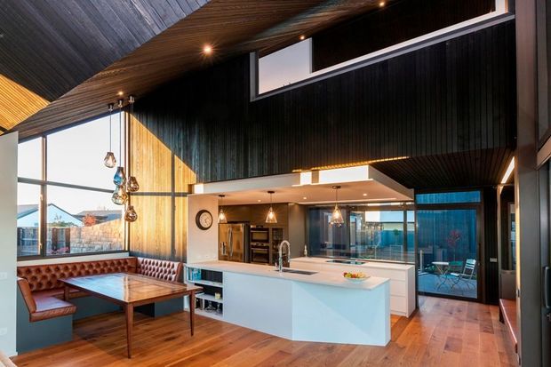 Open-plan contemporary kitchen and dining area with a prominent white island, integrated appliances, dark vertical wall cladding, extensive glazing, and a dramatic vaulted timber-lined ceiling. A built-in tufted leather dining bench and table sit beside the kitchen, while glass doors open to an outdoor area.