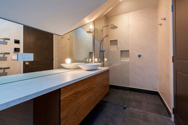 Contemporary bathroom with a floating timber vanity, pale stone-look benchtop, twin vessel basins, large wall mirror, and a walk-in glass shower. The shower has pale textured wall tiles, recessed niches, dual shower fixtures, and dark floor tiles with matching dark skirting. Warm integrated lighting and wood-toned cabinetry create a refined, polished atmosphere.