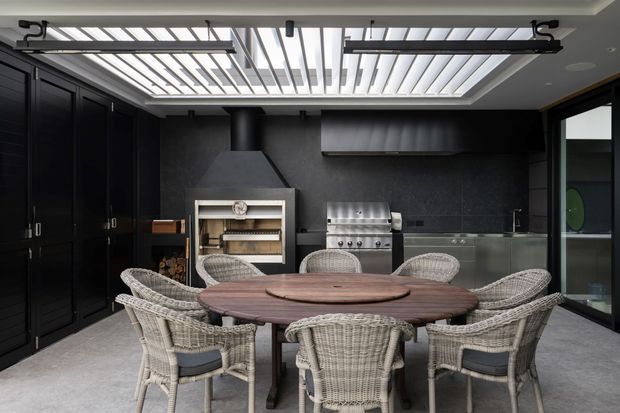A sophisticated covered outdoor kitchen and dining area with a large round timber dining table surrounded by woven wicker chairs. The space features dark built-in cabinetry, a black stone or tile wall finish, stainless-steel barbecue, fireplace or pizza oven, integrated sink, and a dramatic slatted skylight with exposed black framework. Full-height glazing opens to the side, creating a refined indoor-outdoor entertaining space.