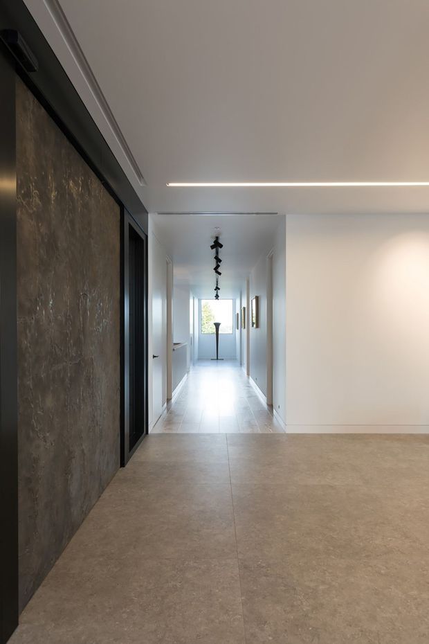 A minimalist contemporary interior hallway and open circulation space with large beige stone or porcelain floor tiles, white walls and ceiling, dark-framed doorway, and a textured dark feature wall. Linear recessed ceiling lighting leads toward a bright glazed end of the corridor, with sculptural black pendant lighting and framed artwork along the hall.