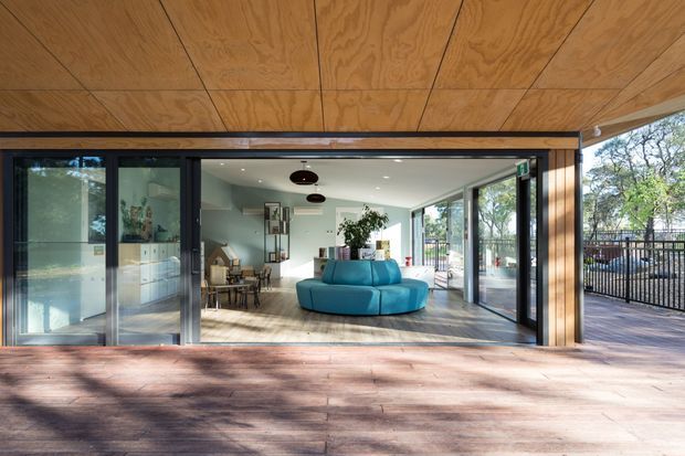 A contemporary open-plan living and dining area viewed from a broad timber deck through fully glazed, dark-framed sliding doors. The interior features a bright turquoise curved sofa, dining table and chairs, light cabinetry, extensive glazing, and a timber-lined ceiling extending over the covered outdoor threshold. A fenced deck and leafy surroundings are visible beyond.