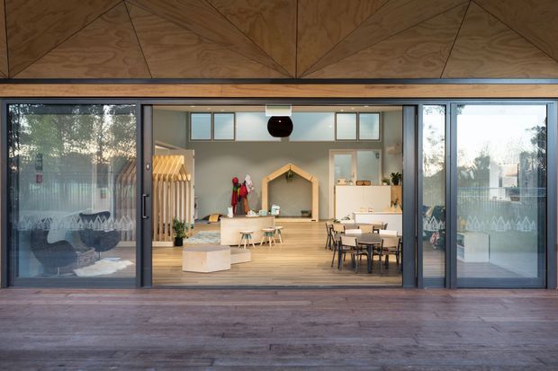 View from a timber deck through wide sliding glazed doors into a bright, open-plan communal interior. The room contains child-sized tables and chairs, soft seating, open shelving, a timber-framed playhouse-like feature, and colourful hanging items. A distinctive geometric plywood-clad ceiling or upper facade is visible above the glazing, with dark-framed doors and warm timber flooring.
