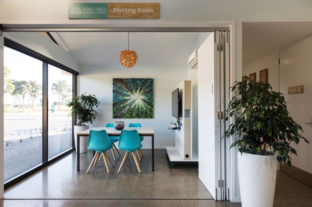 Bright contemporary meeting room with a compact dining-style meeting table and four turquoise chairs, polished concrete flooring, white walls and cabinetry, expansive black-framed glazing, a wall-mounted television, indoor plants, and a sculptural pendant light. The room opens to a hallway through large folding glass doors and appears to be part of a commercial office or shared workspace.