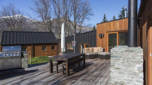 A timber-clad outdoor living deck in a mountainous setting, featuring a dark metal-roofed pavilion, built-in stainless-steel barbecue, outdoor dining table, sectional seating, basketball hoop, and a prominent stone-clad chimney with black flue. The deck and warm timber walls create a rustic contemporary retreat.