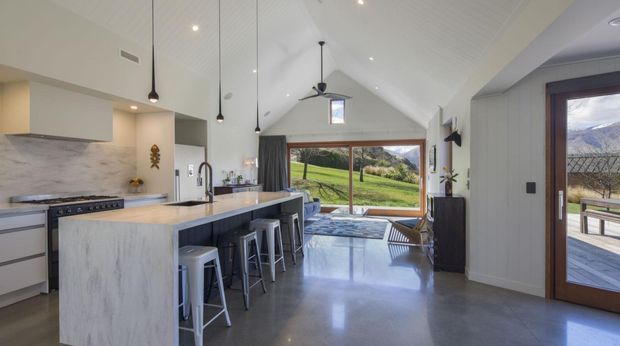 A bright open-plan kitchen and living area with a dramatic vaulted ceiling. The kitchen features a large pale stone island with seating for four, white cabinetry, a matching stone splashback, stainless-steel appliances and three black pendant lights. Polished concrete flooring extends through the space, while full-height glazing and timber-framed doors open to a deck with expansive green rural and mountain views. The interior is clean, contemporary and airy, with a ceiling fan and exposed vaulted form adding architectural character.