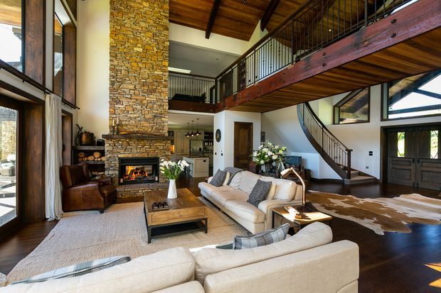 A spacious rustic lodge-style living room with a double-height volume, exposed timber ceiling and beams, a prominent stacked-stone fireplace, large windows, and a mezzanine walkway with black metal balustrades. Two light upholstered sofas, leather lounge chairs, a timber coffee table, and a cowhide rug furnish the warm, open-plan space. A kitchen and stairway are visible beyond the living area.