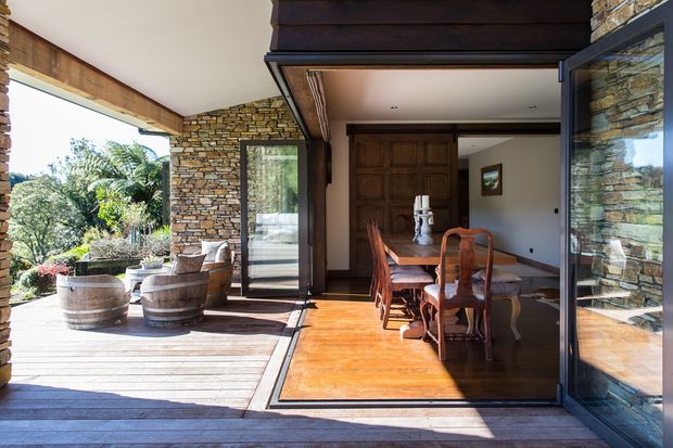 A bright indoor-outdoor dining space with a timber dining table and chairs opening onto a timber deck through large glazed folding or sliding doors. The room features warm polished timber flooring, dark timber ceiling and joinery, and extensive stacked-stone walls and columns. A small outdoor seating area and lush landscaping are visible beyond the deck.