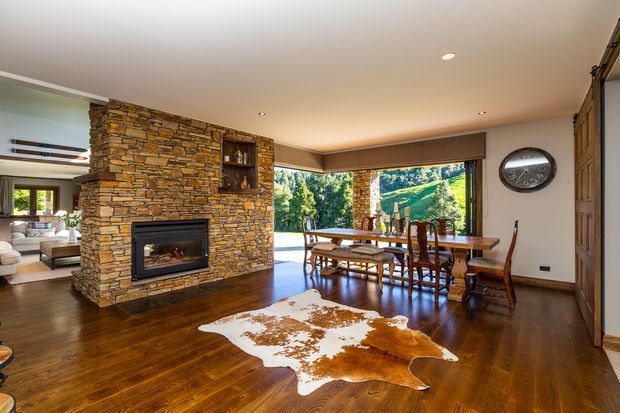 Open-plan dining and living area with a substantial stacked-stone fireplace wall, dark timber flooring, timber dining furniture, and expansive corner glazing overlooking a green landscape. A cowhide rug and exposed timber details add rustic warmth, while recessed lighting and clean-lined window coverings provide a polished contemporary finish.