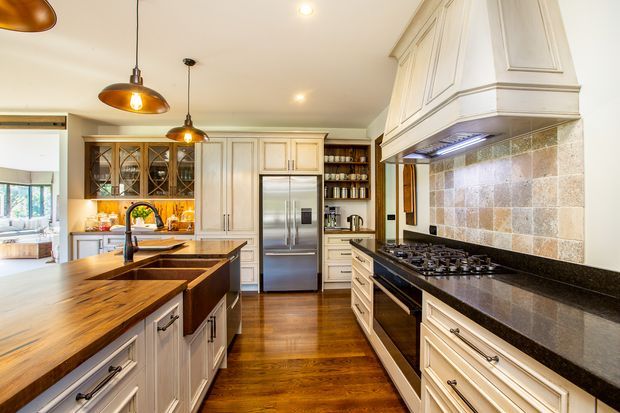 A spacious, traditionally styled kitchen with white paneled cabinetry, glass-front display cupboards, a large timber-topped island, stainless-steel appliances, and a substantial range cooker beneath a decorative hood. The room features warm timber flooring, dark stone benchtops, a multicoloured stone-tile splashback, and pendant lighting.