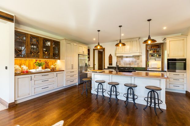 Spacious, warm-toned kitchen featuring extensive cream-painted cabinetry, a large central island with seating for five, dark hardwood flooring, a tiled backsplash, integrated stainless-steel appliances, glass-front display cabinetry, and three pendant lights over the island.